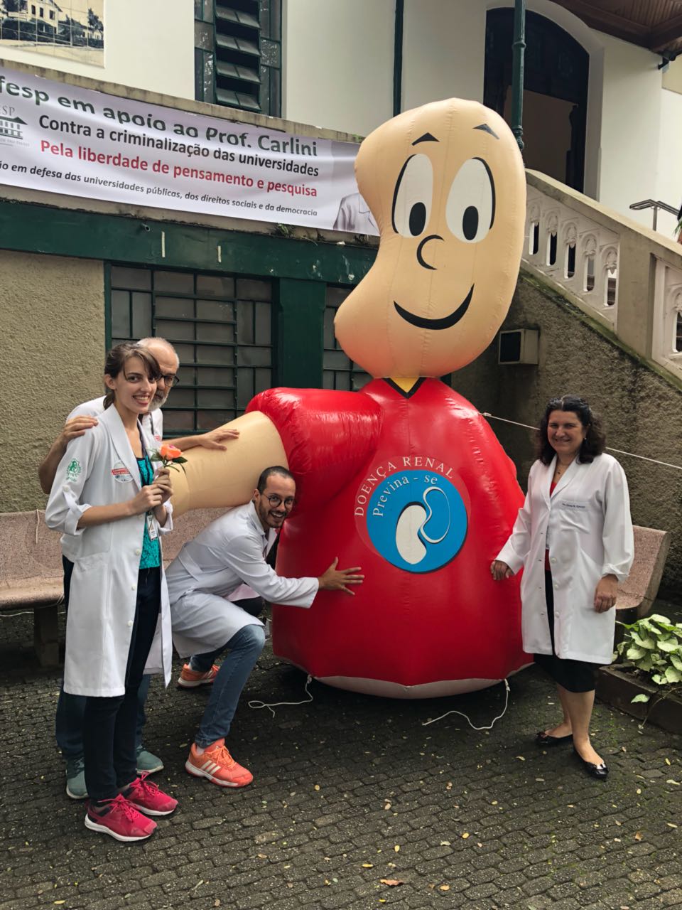 Foto de 4 pessoas vestindo jaleco branco em frente à um boneco vestido de camiseta vermelha escrito "Doença Renal, previna-se"