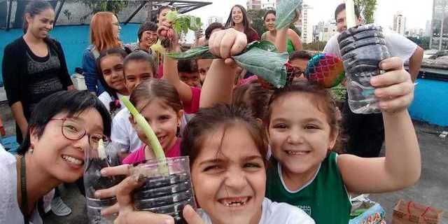 Crianças sorridentes mostram vasos plantados ao fundo adultos observam também sorridentes