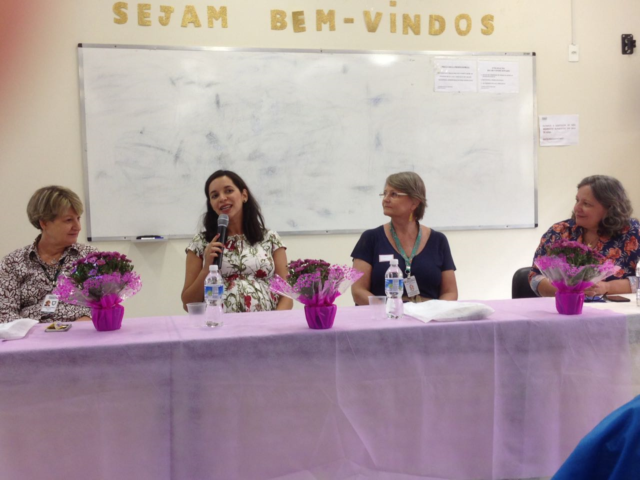 fotografia da diretora da escola paulista de medicina participando da mesa de abertura  do evento. Ao fundo um quadro branco, acima do quadro branco letras formam a frase sejam bem vindos. A mesa está coberta por um pano roxo.